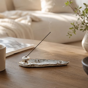 May include: A handcrafted ceramic incense holder with an elongated, irregular leaf-like shape and a white glaze featuring brown textured edges. A small mushroom-shaped stand is attached to one end, holding a thin, lit black incense stick.