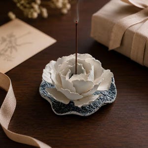 Può includere: Un porta incenso in ceramica bianca a forma di fiore in fiore con petali stratificati, poggiante su una base testurizzata blu scuro. Un sottile bastoncino di incenso marrone scuro si erge verticalmente al centro del fiore.