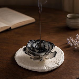 May include: A black and white flower-shaped incense holder sits on a textured white circular base. A thin brown incense stick is placed in the centre of the flower, with a thin trail of smoke rising vertically from the lit tip of the stick.