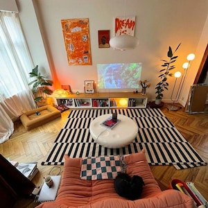 Peut inclure: Un salon confortable à l'esthétique moderne. Il comprend un tapis à rayures noires et blanches, une table basse blanche et un canapé corail. Des œuvres d'art et des plantes ajoutent des touches de couleur. Une télévision est fixée au-dessus d'une étagère basse avec des livres et des objets décoratifs.