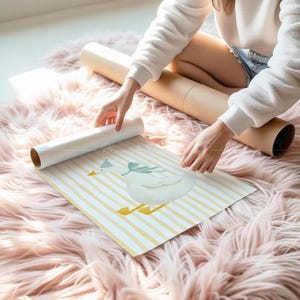 May include: A watercolour print of a white goose wearing a blue bow and hat, on a yellow and white striped background. The print is unrolled on a pink fluffy rug, with a rolled-up print and a brown tube in the background.