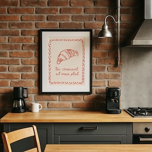 Könnte beinhalten: Ein gerahmter Druck mit einer roten Croissant-Illustration und dem französischen Text "Un croissant s'il vous plaît". Der Druck wird in einer Küche mit einer Backsteinmauer, einer Kaffeemaschine und einer Holz-Arbeitsplatte angezeigt.