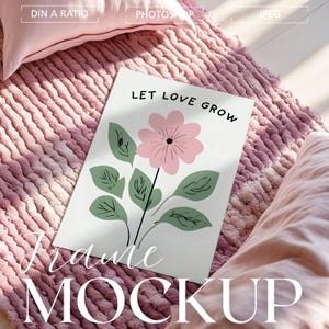 May include: A white framed print featuring the words "LET LOVE GROW" above a pink flower illustration with green leaves. The print rests on a pink textured rug, with a pink pillow in the background. The words "Frame MOCKUP" are at the bottom.