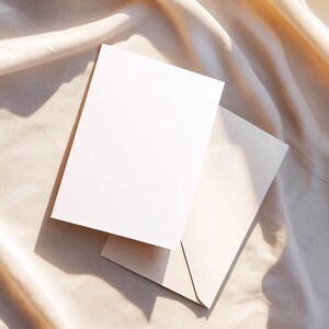 May include: A blank white card and a matching envelope are displayed on a beige fabric surface. The card is rectangular and positioned over the envelope, with both items casting shadows. The scene is lit by natural light, creating a soft, neutral aesthetic.