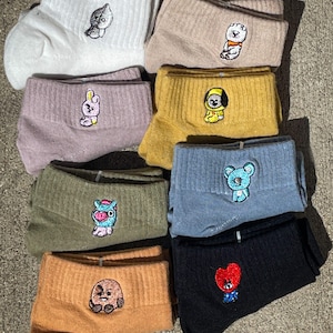 Peut inclure: Collection de chaussettes basses de différentes couleurs : blanc, beige, rose, jaune, vert olive, bleu, orange et noir. Chaque chaussette est ornée d'un personnage de dessin animé brodé. Les chaussettes sont pliées et empilées.