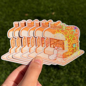 May include: A stack of stickers featuring a cartoon bread shop design. The stickers have a holographic effect and include the text "bread shop" and "bread". The design includes a Shiba Inu dog and a duck.