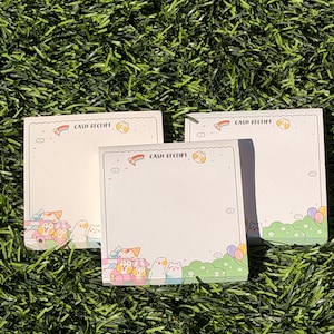 May include: Three square notepads with a cream-colored background and the words "CASH RECEIPT" at the top. Each notepad features a colorful illustration of cartoon animals and a green border with balloons. The notepads are stacked on top of each other.