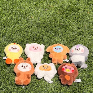 May include: Seven colourful plush toys, each with a unique character design. The toys are in shades of yellow, white, orange, and brown, with embroidered facial features. They are arranged on green grass.