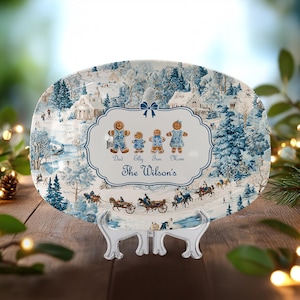 May include: Oval ceramic serving platter with a winter scene in blue and white. The platter features a family of gingerbread people and the name "The Wilson's". The platter is displayed on a clear stand.