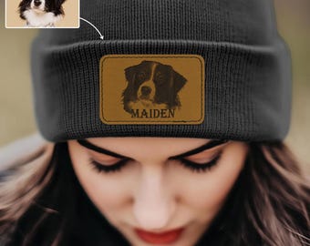 May include: A dark gray beanie with a brown leather patch featuring a dog portrait and the name "MAIDEN". The beanie also has a small image of the dog in the upper left corner.