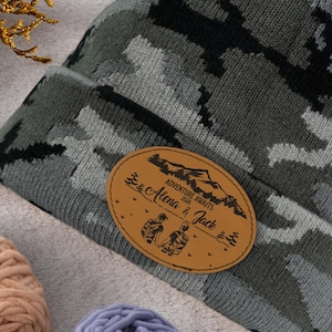 May include: A gray camouflage beanie with a brown leather patch. The patch features the text "Adventure Awaits" and the names "Alena & Jack" with a mountain and tree design. The beanie is made of a soft knit material.