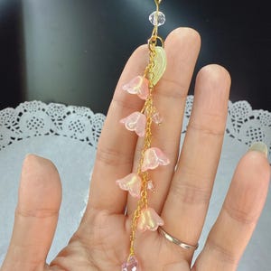 May include: A delicate gold-colored chain with pink flower charms and a clear crystal pendant. The chain is attached to a gold-colored clasp. The flower charms have a translucent appearance.