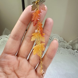 Peut inclure: Ornement suspendu sur le thème de l'automne, avec des feuilles d'érable translucides vertes, oranges et jaunes. Les feuilles sont reliées par des chaînes dorées et ornées de perles de cristal transparentes et ambrées. Un cristal en forme de goutte facettée est suspendu en bas.