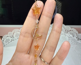 May include: A celestial charm with a gold chain, amber stars, and crystal beads. The charm features a gold crescent moon, a four-pointed star, and clear and amber beads. The charm is suspended by a gold chain, creating a delicate, decorative piece.