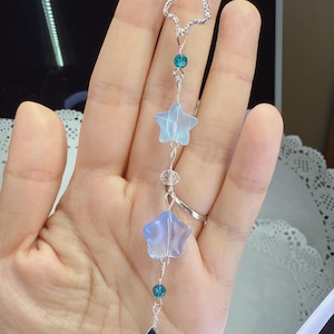 May include: A silver-toned charm necklace featuring a star-shaped outline at the top, with a series of blue star and teardrop-shaped charms. The charms are connected by delicate silver wire and small blue beads. The necklace is held in a hand.