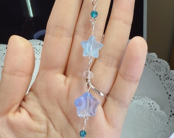 May include: A silver-toned charm necklace featuring a star-shaped outline at the top, with a series of blue star and teardrop-shaped charms. The charms are connected by delicate silver wire and small blue beads. The necklace is held in a hand.