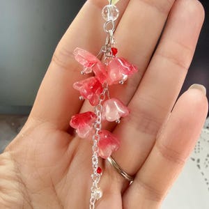 May include: A delicate floral pendant featuring a chain with translucent red and pink flower charms. The pendant includes a yellow leaf-shaped bead, clear crystal beads, and a red teardrop-shaped charm. The jewelry is suspended from a silver chain.