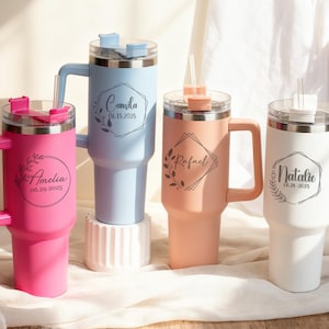May include: Four personalized tumblers in pink, light blue, peach, and white, each with a handle, straw, and lid. The tumblers feature custom names and dates in elegant script within decorative frames. The tumblers are designed for cold beverages.