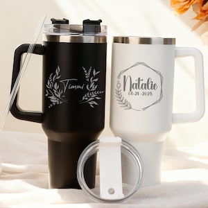 May include: Two personalized travel mugs, one black and one white, with silver rims and handles. The black mug is engraved with "Timmi" and a leaf design. The white mug features "Natalie" and the date 06-28-2025 within a geometric frame.