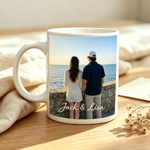 Puede incluir: Una taza de cerámica blanca con una fotografía impresa de dos personas de pie junto a un muro de piedra frente al mar. La imagen muestra a una persona con una blusa blanca texturizada y a otra con camisa azul oscuro y gorra, con el texto Jack & Lisa.