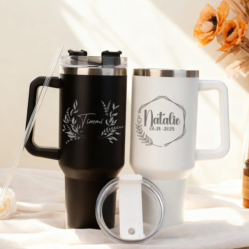 May include: Two personalized travel mugs, one black and one white, with silver rims and handles. The black mug is engraved with "Timmi" and a leaf design. The white mug features "Natalie" and the date 06-28-2025 within a geometric frame.