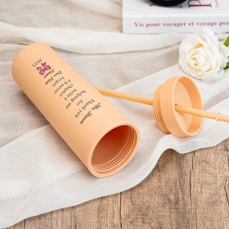 May include: An open peach-colored tumbler with a matching lid and straw. The tumbler has a personalized message and a butterfly graphic. The text reads "Mrs. Brown, Thank you for helping me write a chapter I'll never forget. Love, Emma 2025."