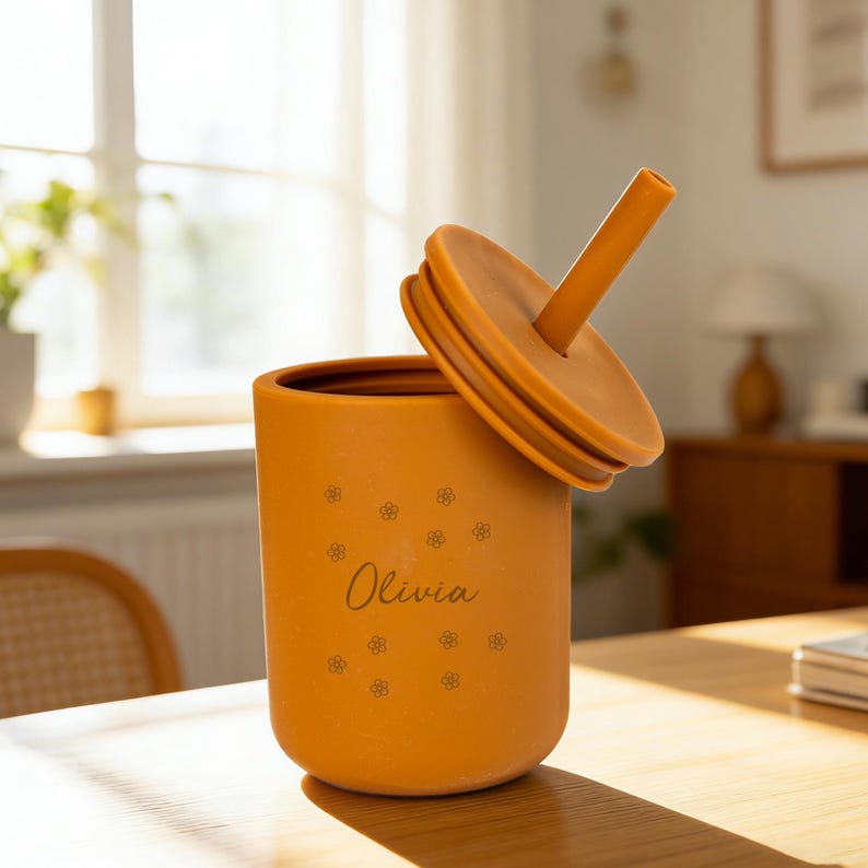 May include: Mustard-colored sippy cup with a matching lid and straw. The cup is decorated with small flower patterns and the name "Olivia" in a cursive font. The lid is slightly ajar, revealing the interior of the cup. The cup is made of a soft material.