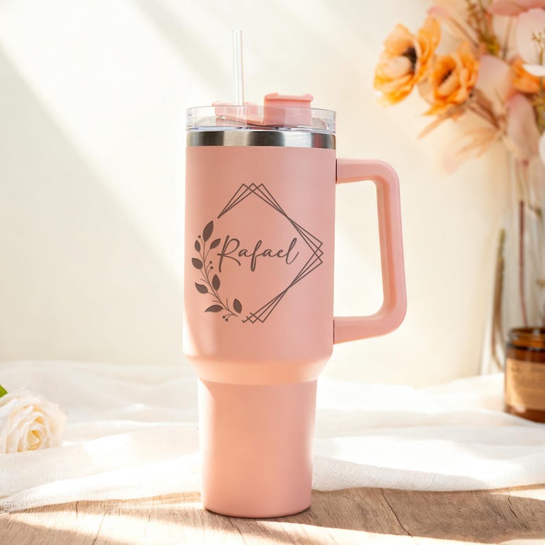 May include: A light pink insulated tumbler with a handle and a clear lid. The tumbler has a straw and a decorative design with the name "Rafael" in a geometric frame. The tumbler is designed to keep beverages cold or hot.