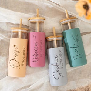 Op de afbeelding: Vier glazen bekers met bamboe deksels en rietjes, elk voorzien van een gekleurde siliconen hoes. De hoezen zijn perzik, roze, lichtgrijs en groenblauw, bedrukt met namen en titels: Denise Bridesmaid, Rebecca Bridesmaid, Sophia Flower Girl en Nicole Bride.