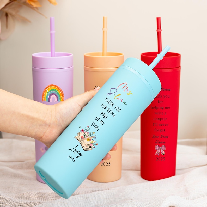 May include: A collection of insulated tumblers in pastel colors, each with a straw. One light blue tumbler features text that reads "Thank you for being part of my story" and "Lucy 2025". Other tumblers are purple, orange, and red.