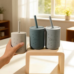 May include: Three silicone cups with lids and straws. One is held by a hand. The cups are light gray and blue-gray, with the names "Harper" and "Olivia" printed on two of them. The cups have a minimalist design with small decorative elements.
