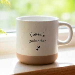 Puede incluir: Una taza de cerámica blanca con acabado moteado y base marrón. La taza tiene las palabras "Vivian's godmother" impresas en escritura negra, junto con dos pequeñas ilustraciones de corazones. La taza está sobre una superficie de madera.