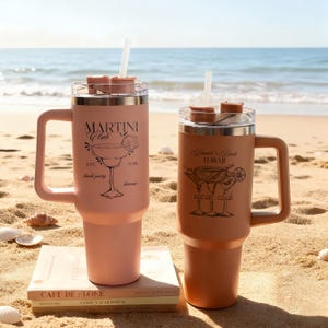 Pu&ograve; includere: Due tazze da viaggio isolate con manici e cannucce su una spiaggia sabbiosa. Una &egrave; color pesca con il testo "MARTINI Club" e una grafica di cocktail. L'altra &egrave; marrone con il testo "Daniel's Beach Hawaii social club" e una grafica di cocktail.