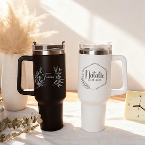 May include: Two personalized travel mugs. One is black with "Timmi" and floral design, the other is white with "Natalie" and date in a geometric frame. Both have handles and clear lids.