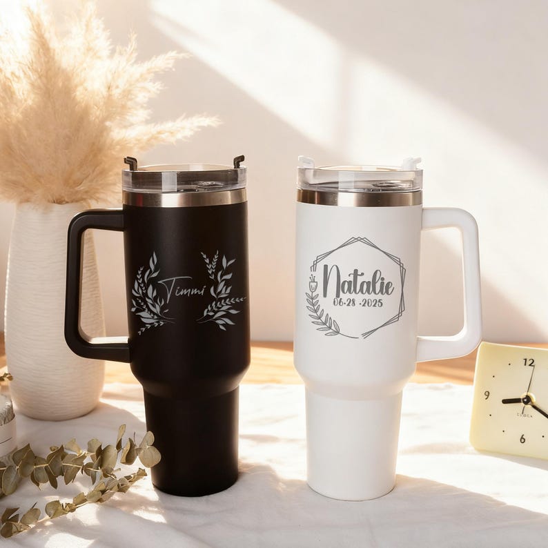 May include: Two personalized travel mugs. One is black with "Timmi" and floral design, the other is white with "Natalie" and date in a geometric frame. Both have handles and clear lids.