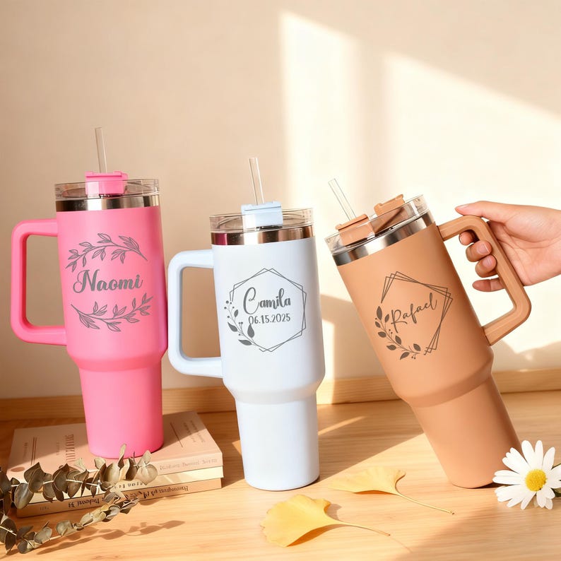 May include: Three personalized insulated tumblers with handles and straws. One is pink with "Naomi" and a floral design, another is white with "Camila 06.15.2025", and the third is tan with "Rafael".