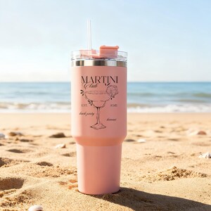 Pu&ograve; includere: Un bicchiere termico rosa con cannuccia e coperchio trasparenti, con la scritta "MARTINI Club" e un'illustrazione di bicchiere da martini. Il bicchiere &egrave; su una spiaggia sabbiosa con l'oceano sullo sfondo. Il testo include anche "Est. 2025 beach party hawaii".