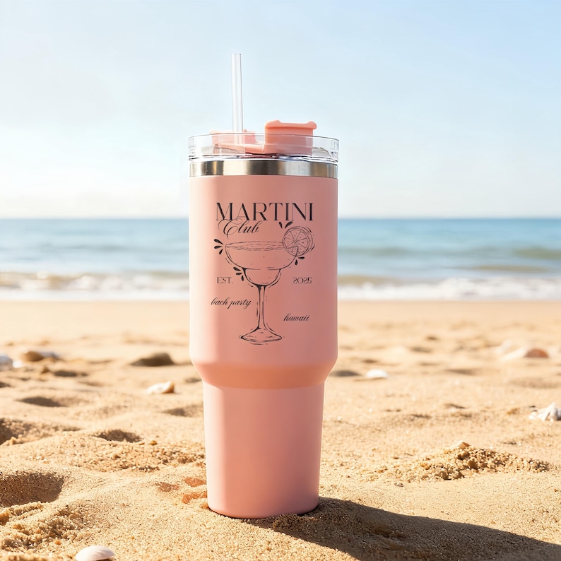 Pu&ograve; includere: Un bicchiere termico rosa con cannuccia e coperchio trasparenti, con la scritta "MARTINI Club" e un'illustrazione di bicchiere da martini. Il bicchiere &egrave; su una spiaggia sabbiosa con l'oceano sullo sfondo. Il testo include anche "Est. 2025 beach party hawaii".