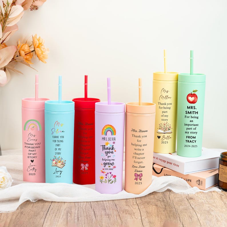 May include: A collection of personalized tumblers with straws in various colors, including pink, blue, red, purple, yellow, and green. Each tumbler features custom text and graphics, such as rainbows, flowers, and the text "Thank you".