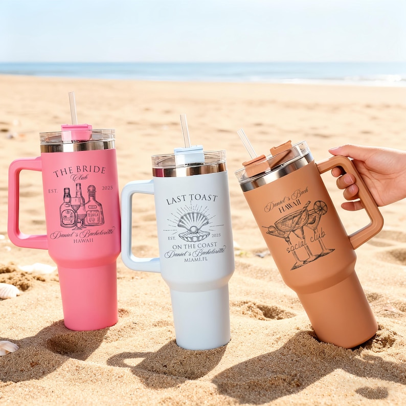 May include: Three insulated travel mugs with handles and straws are displayed on a sandy beach. The mugs are pink, white, and tan, each with a different design and text: "THE BRIDE", "LAST TOAST", and "Daniel's Bach Social Club".