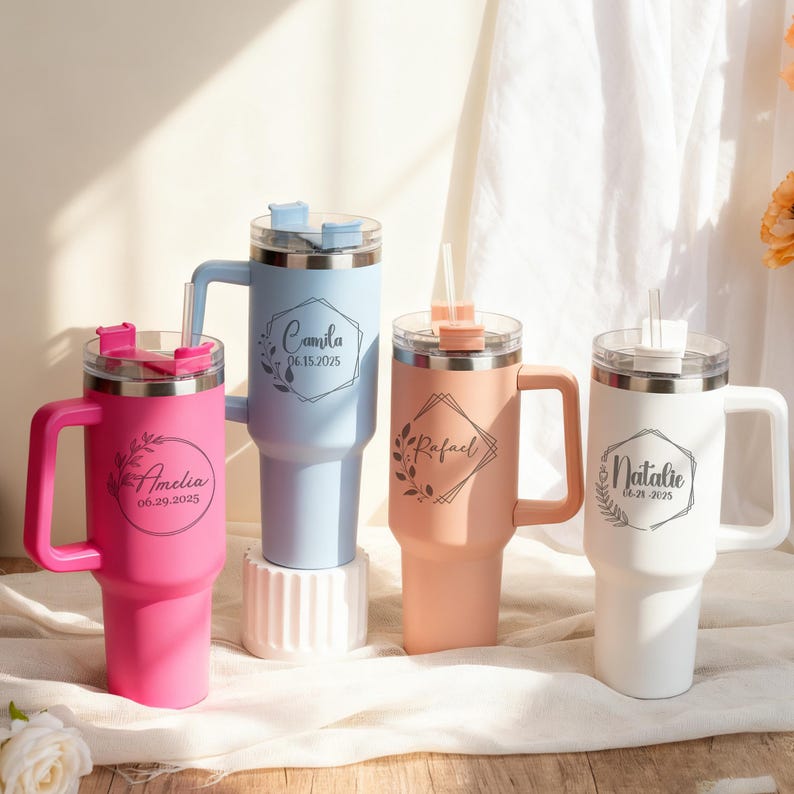 May include: Four personalized tumblers in pink, light blue, peach, and white, each with a handle, straw, and lid. The tumblers feature custom names and dates in elegant script within decorative frames. The tumblers are designed for cold beverages.