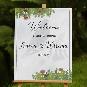 Może przedstawiać: Drewniany sztalug prezentuje tablicę powitalną weselną. Na tablicy widnieje napis "Welcome", imiona "Tracey & Wiremu" oraz data 27.10.2025. Zielone liście i czerwone kwiaty zdobią górę i dół.