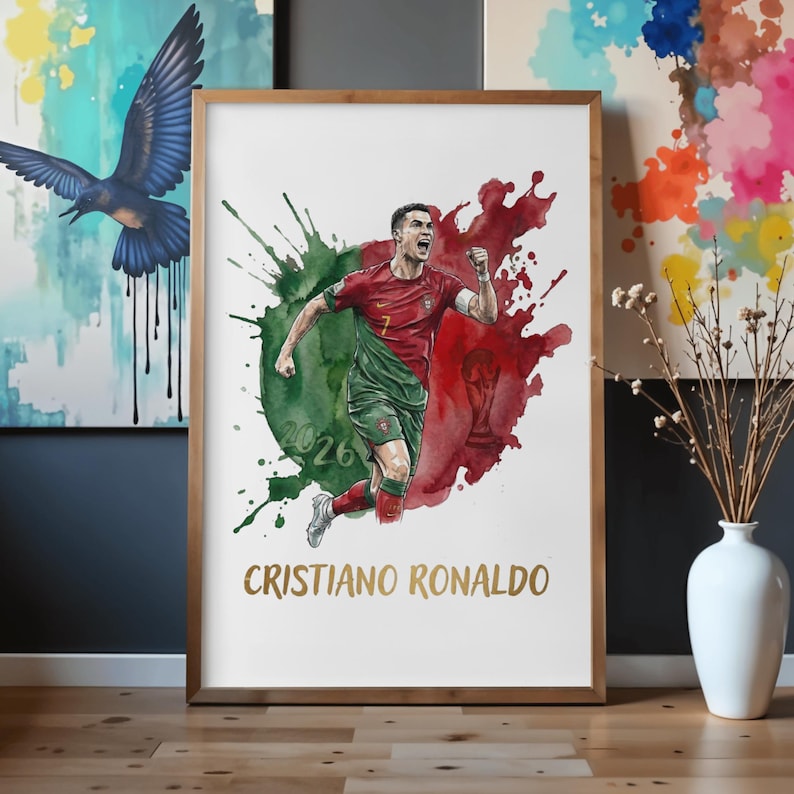 Watercolor style sports poster featuring Cristiano Ronaldo celebrating in a red and green Portugal football kit. The background shows abstract green and red paint splatters mimicking the Portuguese flag, with a subtle World Cup trophy silhouette and the year 2026. The name "CRISTIANO RONALDO" is printed in an elegant gold font at the bottom. The print is showcased in a light wood frame resting on a floor.