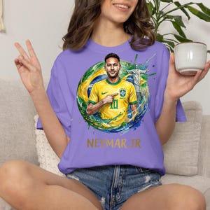 Puede incluir: Una camiseta de color morado claro con una ilustraci&oacute;n gr&aacute;fica de Neymar Jr. vistiendo una camiseta de f&uacute;tbol amarilla de Brasil. El dise&ntilde;o incluye un fondo circular con la bandera brasile&ntilde;a, el Cristo Redentor y el texto NEYMAR JR en dorado.