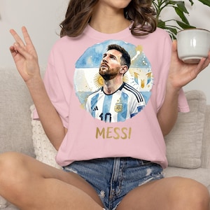 Puede incluir: Una camiseta rosa claro con un gr&aacute;fico circular de estilo acuarela de Lionel Messi vistiendo la camiseta de la selecci&oacute;n argentina. El dise&ntilde;o incluye la bandera argentina, el n&uacute;mero 10 y el texto MESSI impreso en dorado debajo del gr&aacute;fico.