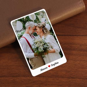 Puede incluir: Una tarjeta fotográfica blanca con una foto de boda de una pareja, con los nombres "Mason" y "Sophia" y un símbolo de corazón. La tarjeta está colocada sobre una cartera de cuero marrón y una superficie de madera.