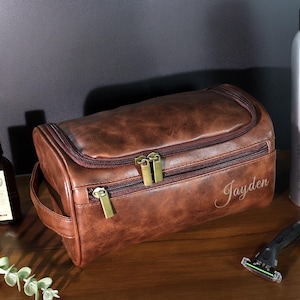 Op de afbeelding: Een bruine leren toilettas met gouden ritsen en de naam "Jayden" staat op de voorkant. Andere items zijn een zilveren fles, een scheermes en twee kleine flesjes. De tas heeft een handvat en een ritssluiting.