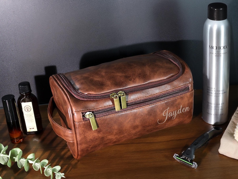 Op de afbeelding: Een bruine leren toilettas met gouden ritsen en de naam "Jayden" staat op de voorkant. Andere items zijn een zilveren fles, een scheermes en twee kleine flesjes. De tas heeft een handvat en een ritssluiting.