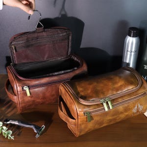 Op de afbeelding: Twee bruine leren toilettassen met gouden ritsen. E&eacute;n tas is open en toont een donkerbruin interieur. De andere tas is gesloten. Andere items zijn flessen met verzorgingsproducten, een scheermes en een handdoek.