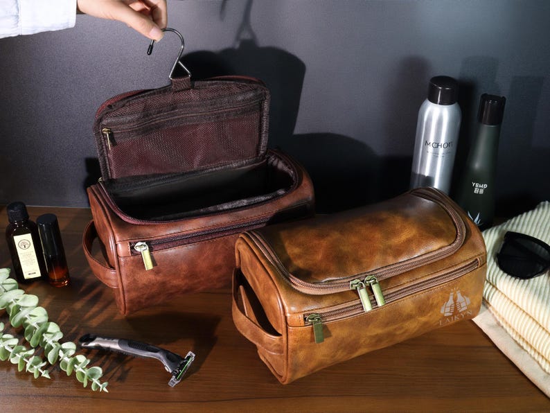 Op de afbeelding: Twee bruine leren toilettassen met gouden ritsen. E&eacute;n tas is open en toont een donkerbruin interieur. De andere tas is gesloten. Andere items zijn flessen met verzorgingsproducten, een scheermes en een handdoek.
